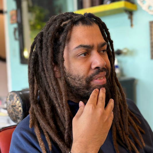 Man with dreadlocks sitting in a chair, wearing a watch, in a barbershop setting.