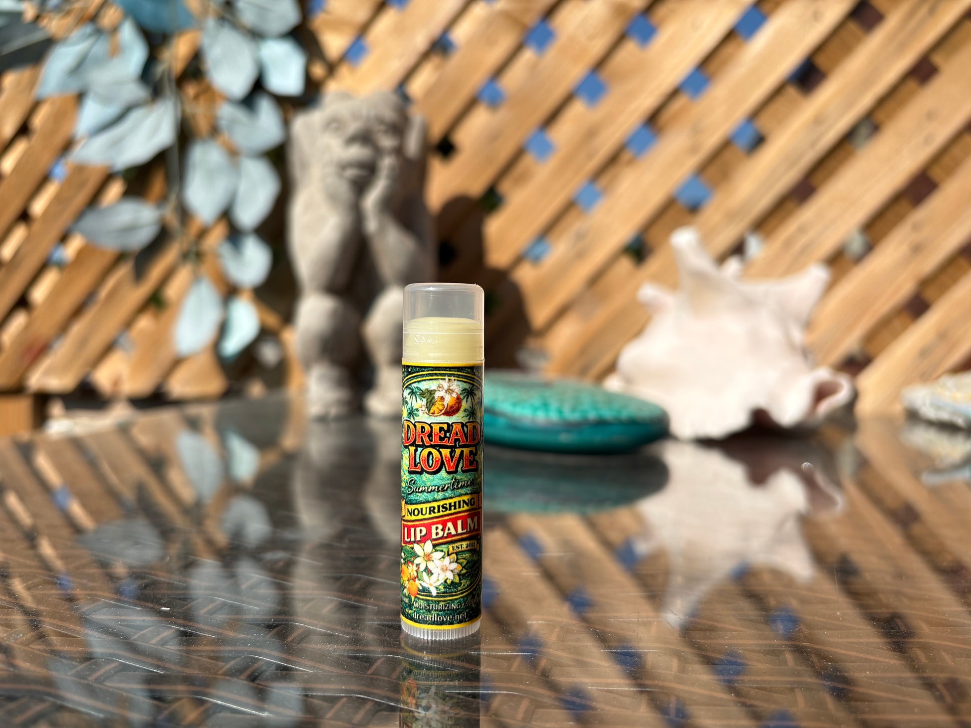 Dread Love Summertime Creamsicle Vegan Nourishing Lip Balm tube on an outdoor glass table with a garden gnome and seashell backdrop