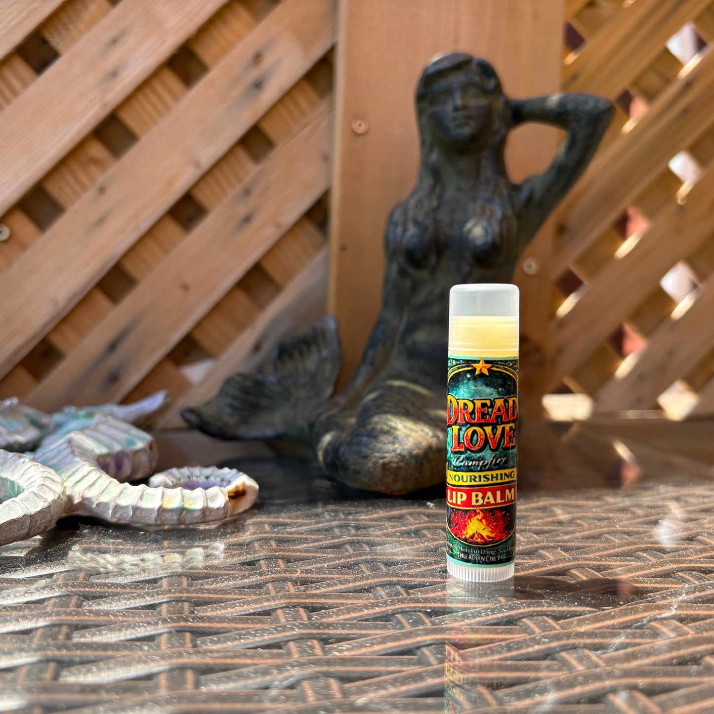 A tube of Dread Love Lip Balm stands on a textured metal patio table in front of a wooden lattice fence, with a bronze mermaid statue and a decorative snake sculpture in the background.