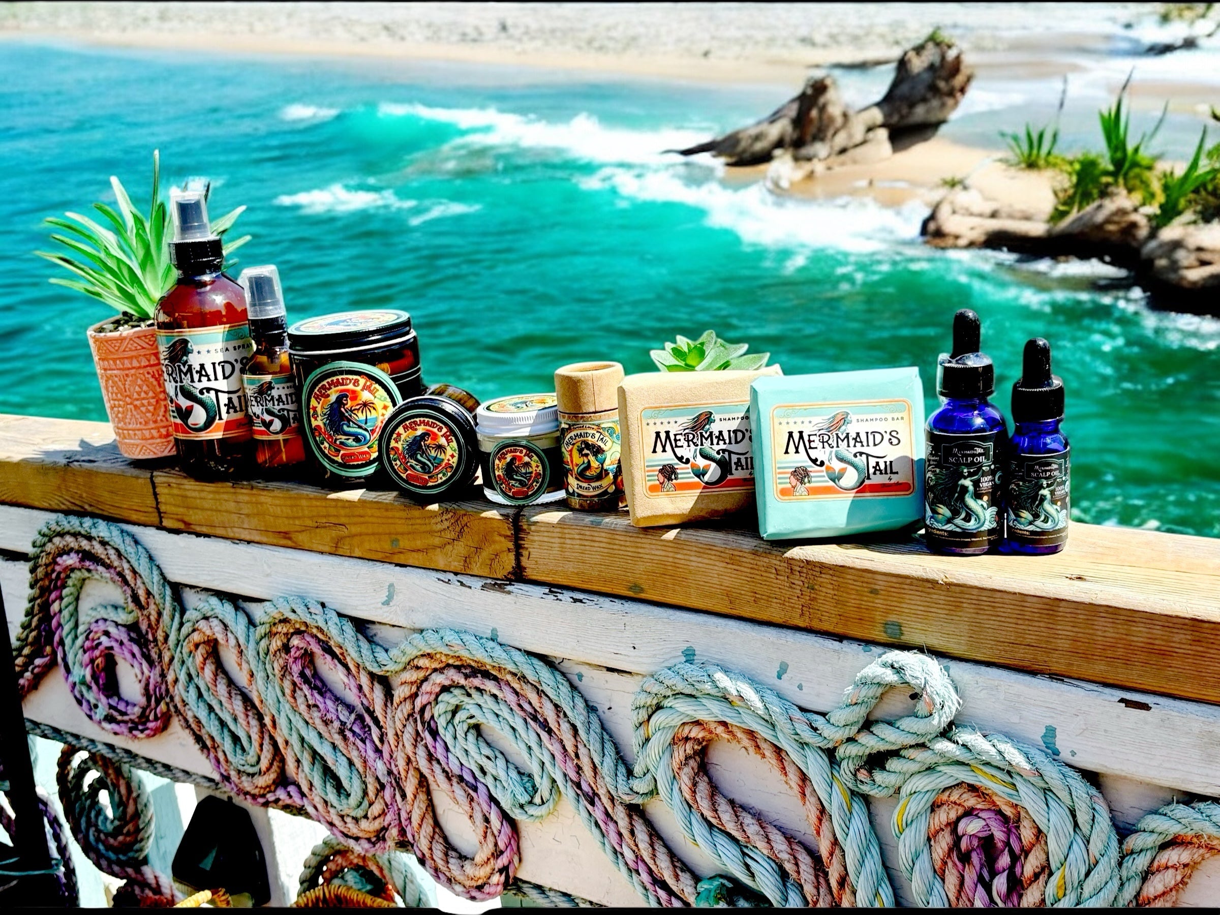Handmade Dread Love Mermaid's Tail dreadlock care products displayed on a wooden railing by the ocean, including sprays, scalp oils and styling wax