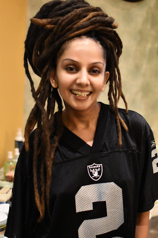Person long dreadlocks and a gold grill wearing a black sports jersey with a logo and number, standing indoors.