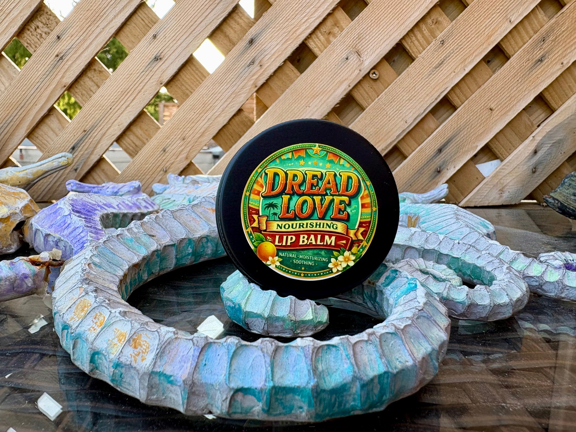 A round black tin of Dread Love Summerime Vegan Nourishing Lip Balm sits on top of a large coiled blue and white sea urchin shell structure in front of a wooden lattice fence.