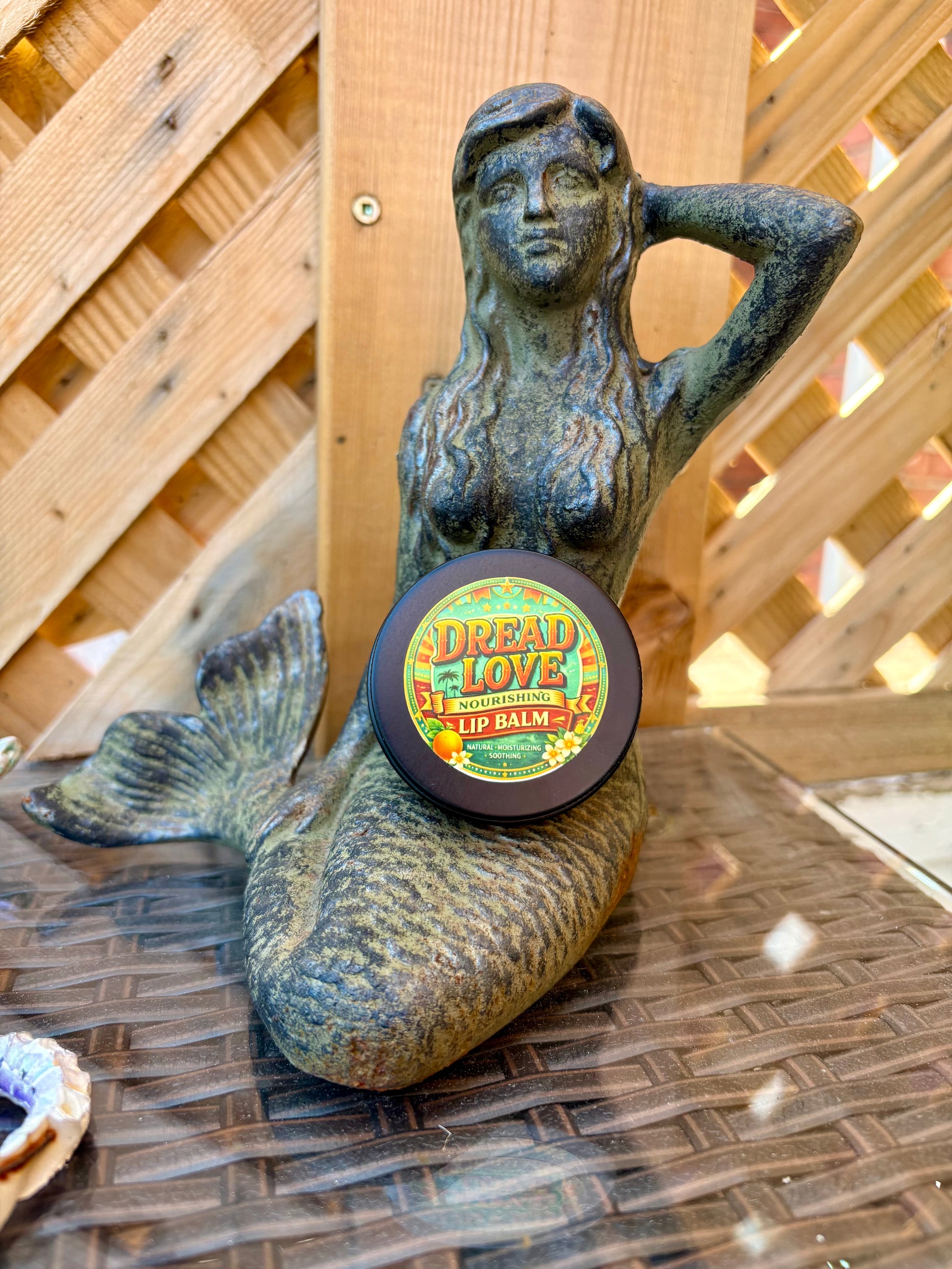 A round tin of Summertime lip balm by  Dread Love is displayed on a bronze mermaid statue. The product features a colorful retro-style label with yellow, green, and red text.